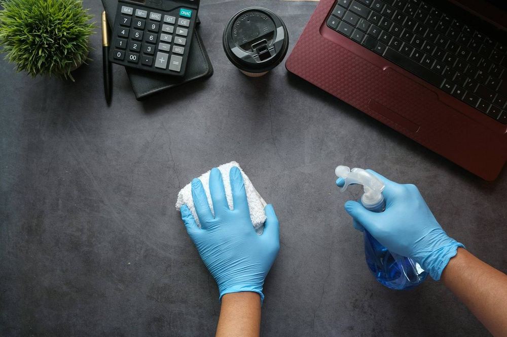 Gloved hands wiping a desk and holding a spray bottle beside a laptop.