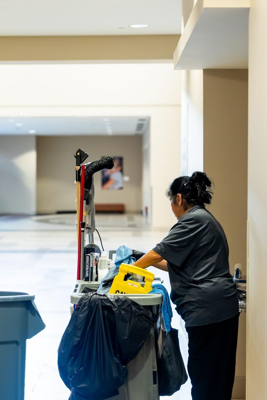A janitor in a gray shirt organizing supplies on a cart in a commercial hallway.